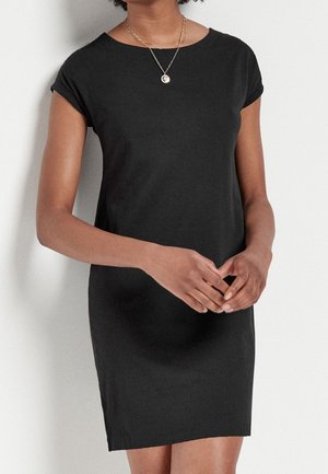 Femme portant une robe noire ajustée à manches courtes et un collier pendentif en argent, les mains doucement jointes devant elle.