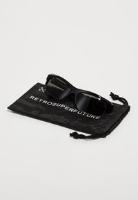 Lunettes de soleil noires avec une monture épaisse et angulaire posées sur un sac à cordon noir avec le texte "RETROSUPERFUTURE" en blanc.