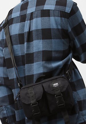 Person trägt ein blau-schwarz kariertes Hemd und eine schwarze Vans-Umhängetasche, die über den Rücken getragen wird.