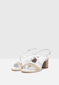 Sandales blanches et beiges avec un talon bloc, des lanières croisées, des accents métalliques et une bride de cheville ajustable. Texture en cuir lisse.