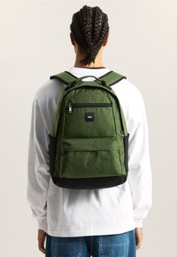 Persona con cabello trenzado que lleva una camisa blanca de manga larga y jeans azules, cargando una mochila Vans verde y negra, de espaldas.