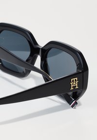Black oversized sunglasses with a hexagonal frame, dark lenses, and gold logo accent on the arm; sleek, smooth finish.