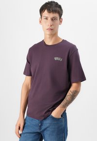 Camiseta de algodón de color púrpura oscuro con mangas cortas, escote redondo y un pequeño logo decorativo en verde en el lado izquierdo del pecho.