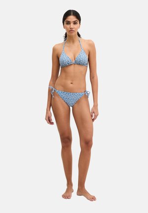 Młoda kobieta stojąca boso, ubrana w niebieski kwiatowy bikini z wiązaniem na szyi i wiązanymi bokami, na jasnym, jednolitym tle.
