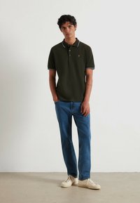 Polo shirt verde scuro con bordo bianco, indossato con jeans azzurri chiari e sneakers beige. Il polo presenta un piccolo logo ricamato sul petto.