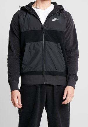 Osoba ubrana w czarną bluzę Nike z kapturem na zamek i przednimi kieszeniami oraz czarne spodnie na jasnym, jednolitym tle.