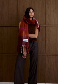 Wollen sjaal met een verloop van tinten rood, roze en paars, met franjes aan de uiteinden en een wit label met de tekst "ARCTIC FOX." Model draagt een bruine top en grijze broek.