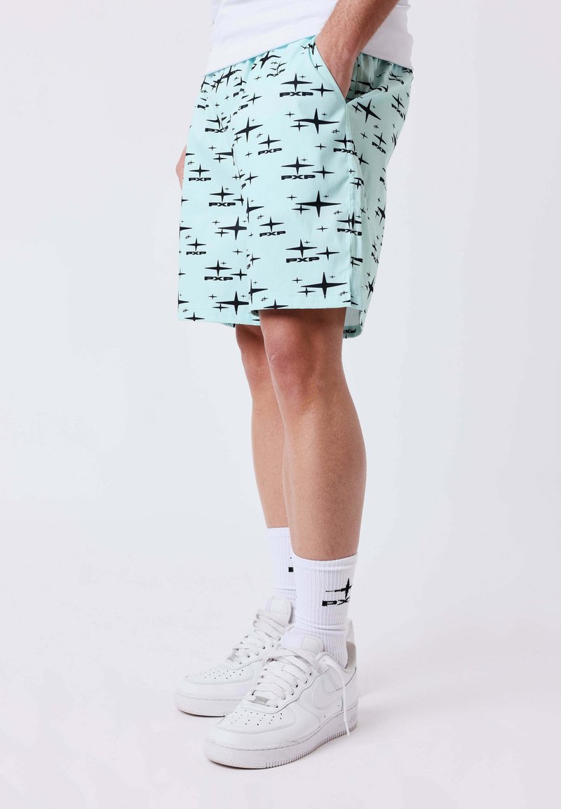 Homme portant un short bleu clair avec des motifs noirs en forme d'étoiles, des chaussettes blanches et des baskets blanches, debout avec une main dans la poche.