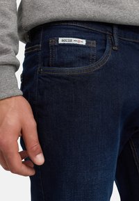 Nærbilde av en persons hånd nær en lomme på mørkeblå denimjeans med synlig etikett "INDICODE siden 1993", iført en grå genser.