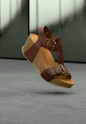 Brun skinn kilehæl sandal med to gullspenner og korksåle som svever over et grått gulv mot en uskarp bakgrunn.