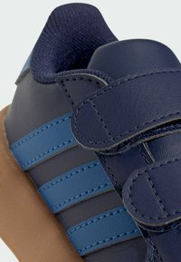 Chaussure en velcro de couleur marine et bleue avec un matériau synthétique lisse, une semelle en caoutchouc marron et un col de cheville texturé. Présente trois rayures bleues.