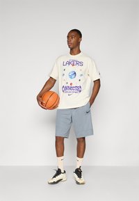 Lys beige t-skjorte med "Lakers" grafikk med planeter, kombinert med grå shorts og svarte/hvite joggesko, holder en oransje basketball.