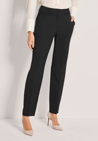 Schwarze maßgeschneiderte Hose mit geradem Bein, mit Standardbund, Seitentaschen und Bundfalten; getragen mit beigen Absätzen.