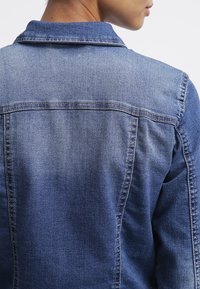 Džínová bunda ve středně modré barvě s klasickým límcem. Má viditelné švy, upravený zadní díl a mírně vybledlou texturu.
