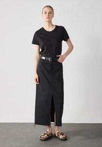 T-shirt noir en coton associé à une jupe en jean noire taille haute avec une fente à l'avant, complété par des sandales à glissière marron à motifs.