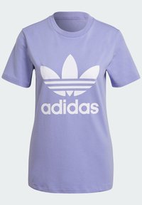 Lila bomull t-shirt med en stor vit Adidas-logotyp och tre ränder under, korta ärmar och rund halsringning.