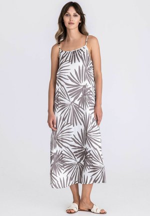 Vrouw die een witte slipjurk draagt met een grijze palmbladprint, dunne bandjes en witte platte sandalen, staand tegen een effen achtergrond.