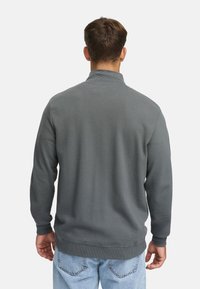 Hombre con cabello corto castaño, vistiendo un suéter gris acanalado y jeans azul claro, de pie con su espalda vuelta hacia un fondo blanco.