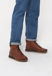 Brązowe skórzane botki sięgające kostki z teksturowanym wykończeniem, sznurowanym fasonem i gumową podeszwą. Noszone z podwiniętymi niebieskimi dżinsami.