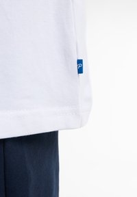 Tricou alb din bumbac, cu o etichetă albastră pe lateral, având un tiv drept și o textură netedă și moale. Material de culoare albastru marin dedesubt.