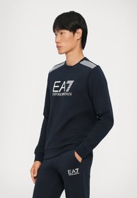 Felpa blu navy con maniche lunghe, collo a giro, dettagli a righe contrastanti sulle spalle e logo EA7 bianco sul davanti.