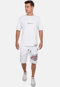Witte katoenen T-shirt met een zwart geborduurd logo, gecombineerd met witte shorts met een rode grafische print. Witte sneakers met rode accenten.