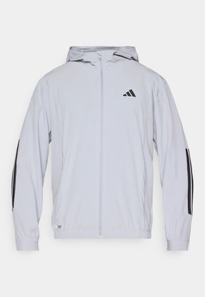 Giacca sportiva grigio chiaro con zip frontale, cappuccio e strisce nere sulle maniche. Realizzata in materiale liscio e leggero. Presenta una fascia in vita aderente.