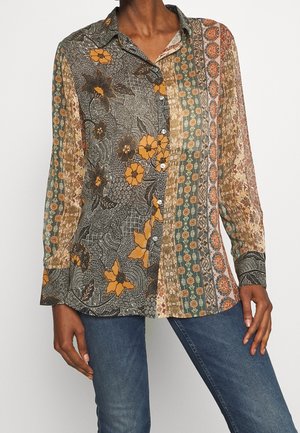 Kvinde iført flerfarvet patchwork blomstret bluse med knapper og lange ærmer samt blå jeans, stående mod en ensfarvet baggrund.