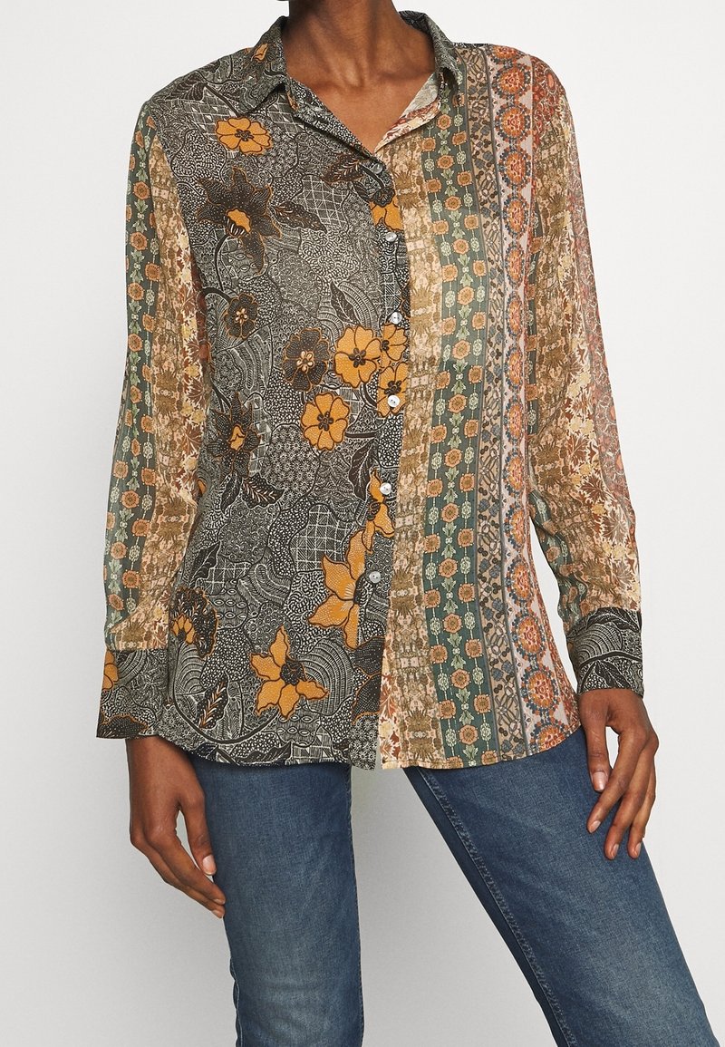 Femme portant un chemisier patchwork fleuri multicolore boutonné à manches longues et un jean bleu, debout devant un fond uni.