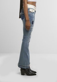 Jeans évasés bleus avec des détails déchirés et des patchs contrastants, dotés d'une taille haute et d'ourlets effilochés, associés à des bottines noires.