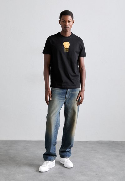 Ein Mann steht gegen eine schlichte Wand und trägt ein schwarzes Versace-T-Shirt mit goldenen Logo, verblasste blaue Jeans und weiße Turnschuhe auf einem Betonboden.