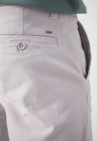 Gros plan sur un pantalon gris clair avec une poche arrière boutonnée et une poche latérale porté avec une chemise vert foncé.
