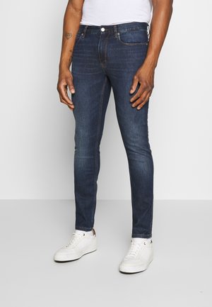 Man die donkerblauwe slim-fit jeans, witte sneakers en een wit T-shirt draagt, staand tegen een effen lichtgrijze achtergrond.