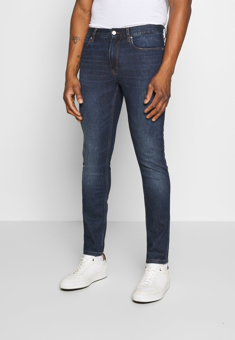 Homme portant un jean slim bleu foncé, des baskets blanches et un t-shirt blanc, debout devant un fond uni gris clair.