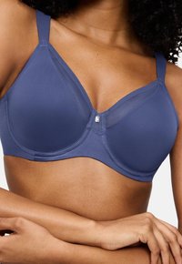 Zblizu prikaz ženske, ki nosi brezšivni modri nedrček z žično oporo, z mrežastimi detajli in prekrižanimi rokami, ki počivajo na pasu.