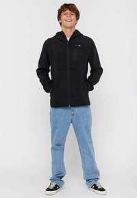 Sort lynlåshættejakke i blød stof med justerbar hætte, frontlommer og en brystlomme. Sammen med lyseblå denim jeans og sneakers.