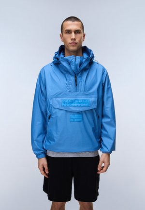 Mann trägt blaue Kapuzen-Windjacke mit Fronttasche und schwarze Shorts vor schlichtem grauem Hintergrund.