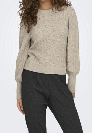 Femme portant un pull en tricot beige à motif ondulé avec de longues manches bouffantes, associé à un pantalon gris foncé texturé.