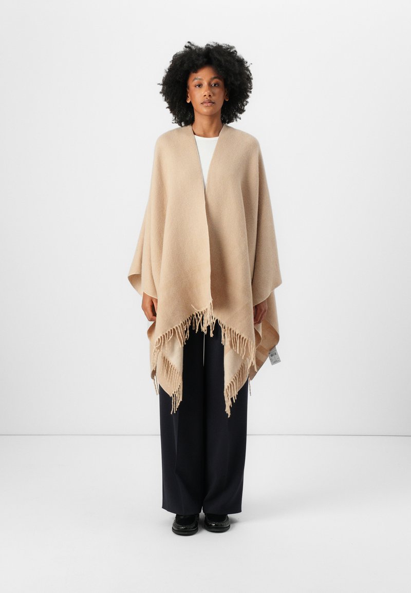 Beige ullponcho med sjalkrage og frynser, som henger løst over en hvit topp og mørke bukser. Glatt tekstur.