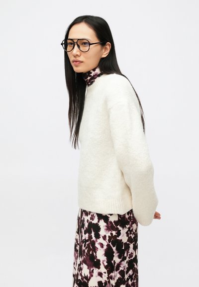 Biely, nadýchaný sveter s vysokým golierom v kombinácii s dlhou vzorovanou sukňou v odtieňoch fialovej a čiernej. Priehľadné okrúhle okuliare.