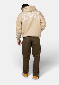 Felpa beige con il testo bianco "CRYSTALPARIS" sulla schiena, abbinata a pantaloni marrone scuro e sneakers con accenti gialli. Look casual, materiale morbido.
