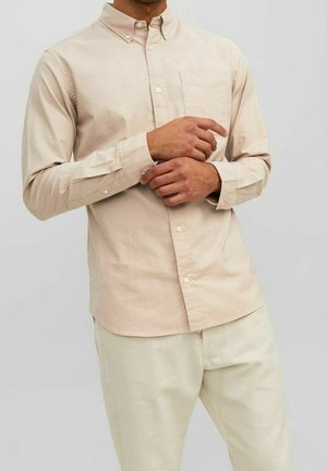 Homme portant une chemise beige à boutons, ajustant sa manche gauche, associée à un pantalon beige clair sur un fond clair uni.
