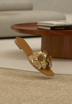 Sandale à bride marron avec grande décoration florale métallique dorée, posée sur un tapis tissé beige devant une table basse en bois et un canapé crème.