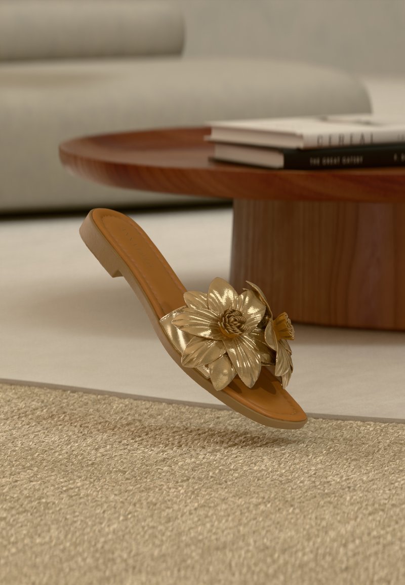 Καφέ πλατφόρμα με μεγάλο μεταλλικό χρυσό λουλούδι, τοποθετημένη σε μπεζ υφαντό χαλί μπροστά από ξύλινο τραπεζάκι καφέ και κρεμ καναπέ.