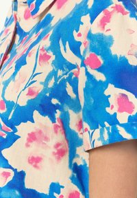 Camicia a maniche corte realizzata in tessuto con uno sfondo blu e motivi floreali rosa. Il colletto è strutturato e il tessuto ha una trama liscia.