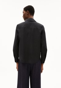 Black long-sleeve shirt with a classic collar, smooth texture, and a slightly curved hem, worn with navy pinstriped pants.