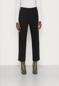Persoon met zwarte rechte broek, witte lange mouw top en olijfgroene gestructureerde enkellaarsjes, staand op een lichte vloer.