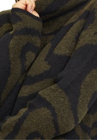 Main remontant le col d'un pull en maille épais, doux, vert foncé et noir à motif, à manches longues.