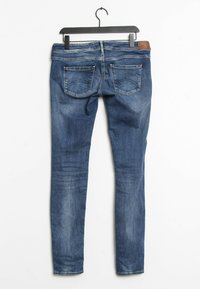 Slim fit -tyyliset siniset farkut, joissa on sileä pinta, viiden taskun malli ja ruskea nahkainen vyötärökoriste. Ripustettu mustaan koukkuun.