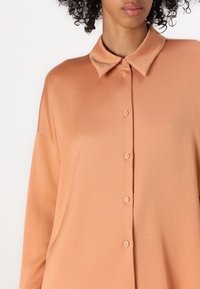 Camisa de botón de color melocotón con cuello clásico, mangas largas y una textura suave, con botones a juego en la parte delantera.